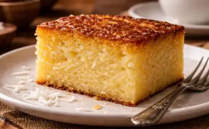 Fatia de bolo de aipim cremoso servida em prato de sobremesa, mostrando a textura úmida e pedaços de coco fresco, acompanhada de uma xícara de café.