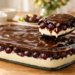 Travessa de vidro transparente mostrando as camadas da sobremesa: creme branco embaixo, muitas uvas roxas Vitória no meio e cobertura de chocolate brilhante por cima, com uma colher servindo.
