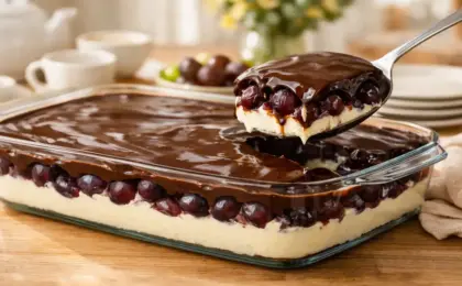 Travessa de vidro transparente mostrando as camadas da sobremesa: creme branco embaixo, muitas uvas roxas Vitória no meio e cobertura de chocolate brilhante por cima, com uma colher servindo.