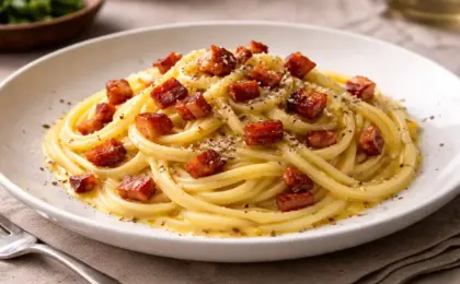 Prato de cerâmica branco com espaguete de palmito pupunha envolto em molho carbonara amarelo cremoso, com pedaços de bacon crocante e pimenta preta por cima.