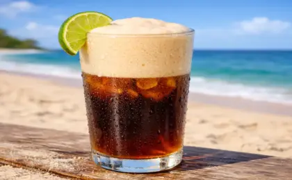 Copo grande de vidro suado com mate gelado escuro e uma camada grossa de espuma de limão bege por cima, decorado com fatia de limão, em cenário de praia.