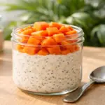 "Foto em close de um pote de vidro transparente contendo Overnight Oats (aveia dormida) em camadas cremosas intercaladas com sementes de chia e cubos frescos de mamão papaia no topo, iluminado por luz natural de manhã."