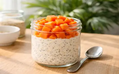 "Foto em close de um pote de vidro transparente contendo Overnight Oats (aveia dormida) em camadas cremosas intercaladas com sementes de chia e cubos frescos de mamão papaia no topo, iluminado por luz natural de manhã."